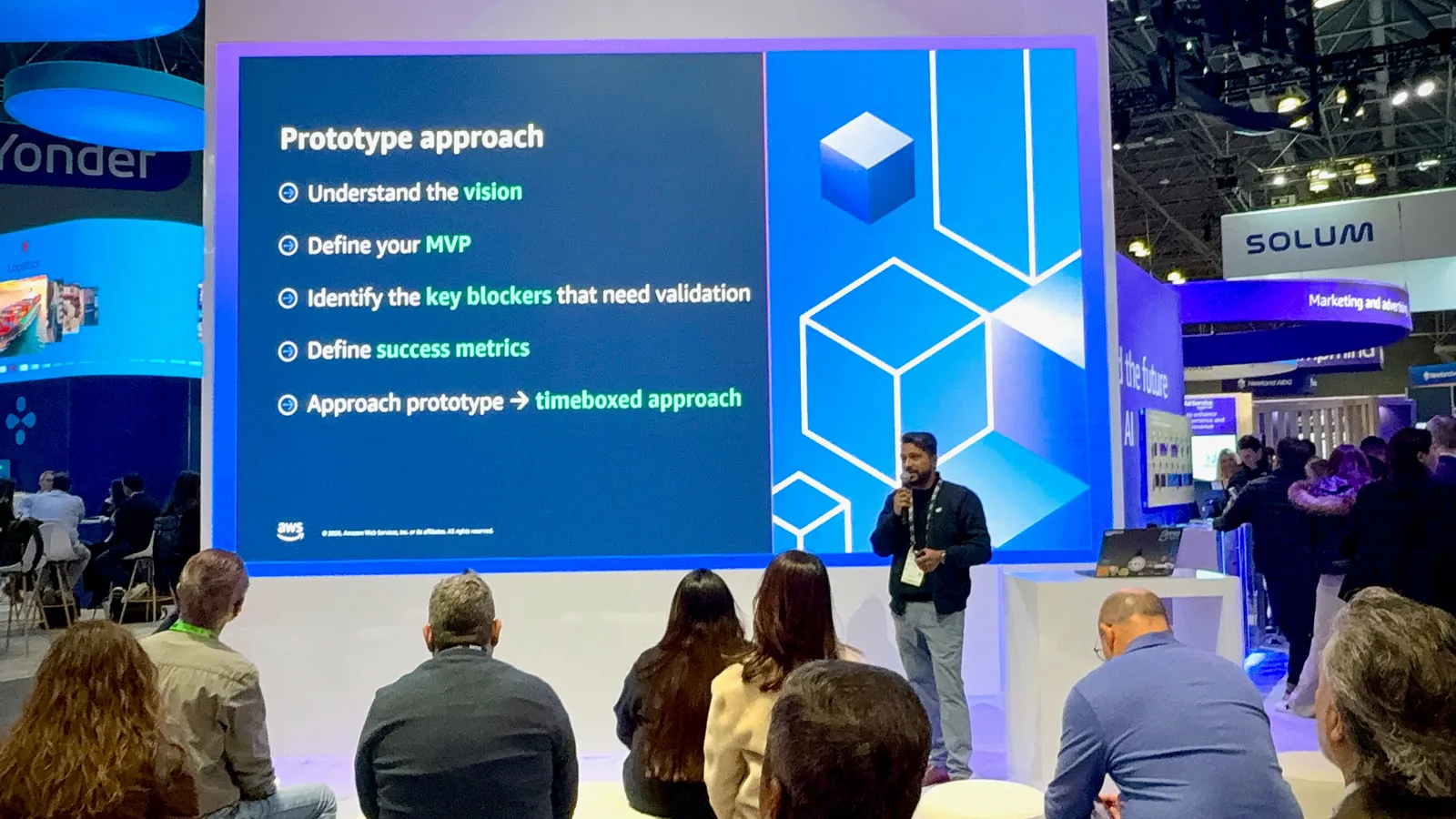 Ganesh Raam on stage at the AWS booth theater, beside a slide titled Prototype approach